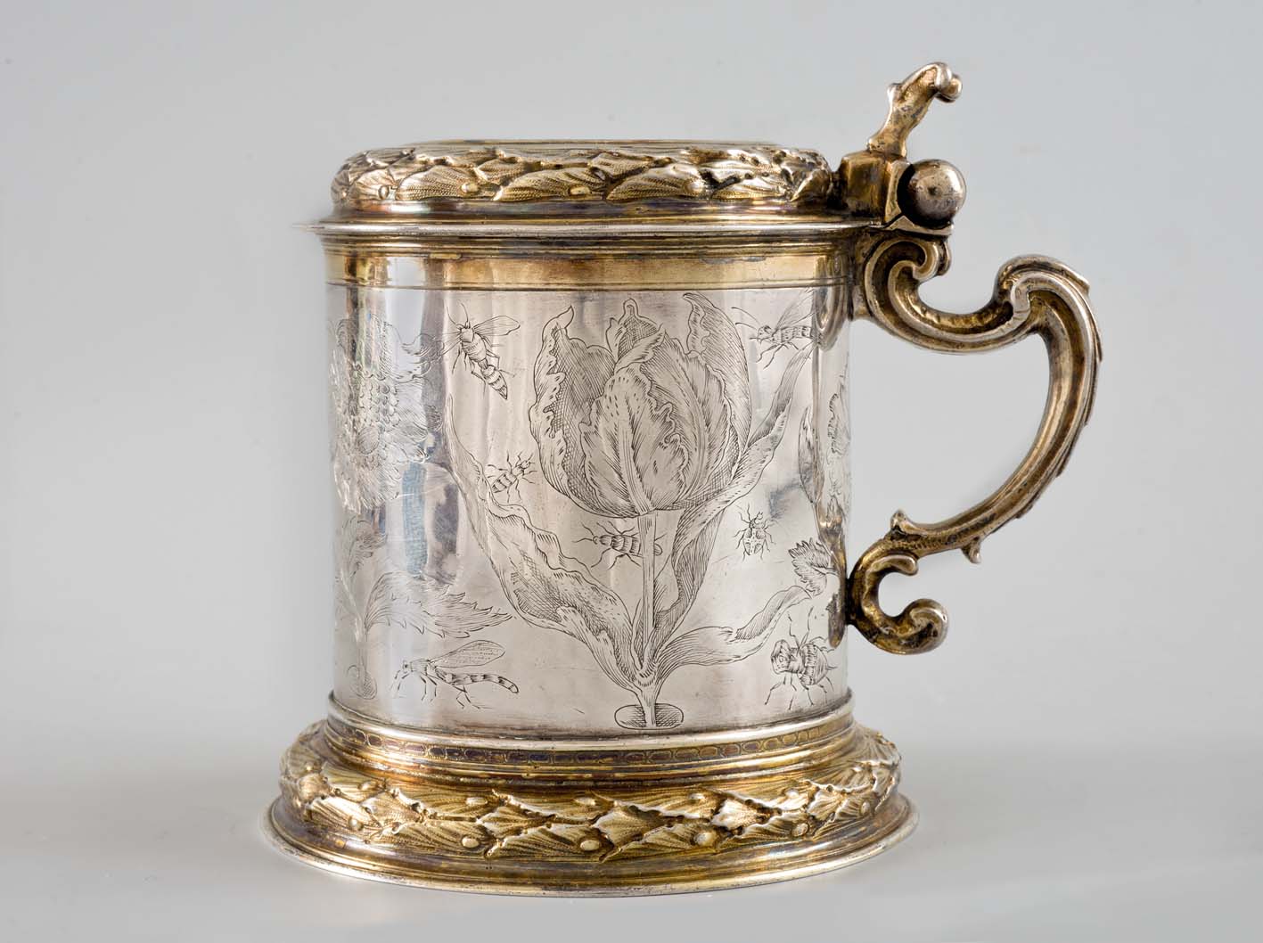 Srebrny kufel ze złotymi motywami zdobiony grawerunkiem. Na jego brzuścu znajdują się kwiaty oraz owady. 
