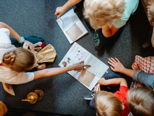Widok z góry na podłogę na której siedzą małe dzieci. Przed nimi kartki z zadaniami edukacyjnymi. Kobieta siedząca po lewej stronie wskazuje swoją ręką na zadanie na kartce.