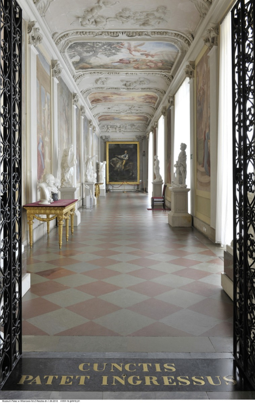 Kolorowe zdjęcie. Przedstawia otwartą bramę i korytarz w pałacu. Na pierwszym planie posadzka kamienna z dwóch kolorów tafli. Po bokach korytarza, na postumentach rzeźby. Na końcu korytarza duży obraz z przedstawieniem mężczyzny na koniu. Na sklepieniu sztukaterie i malowidła. 