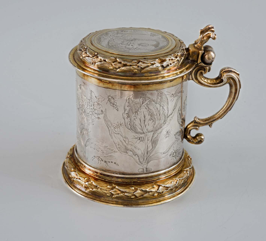 Srebrny kufel ze złotymi motywami zdobiony grawerunkiem. Na jego brzuścu znajdują się kwiaty oraz owady. 
