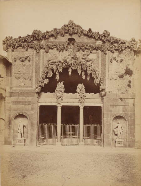 Stara fotografia w kolorze sepii. Przedstawia widok na fasadę budynku antycznego. W dolnej części budynku dwie nisze i dwie rzeźby antyczne: kobiety i mężczyzny. Na środku dwie kolumny. W górnej części fasady otwór zdobiony roślinami. Jest to Grotta Grande.